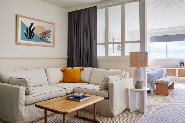 A cozy living space with a cream sofa, a wooden coffee table, and a lamp. There's artwork on the wall and a glimpse of a bedroom.