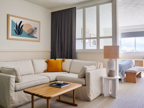 A cozy living space with a cream sofa, a wooden coffee table, and a lamp. There's artwork on the wall and a glimpse of a bedroom.