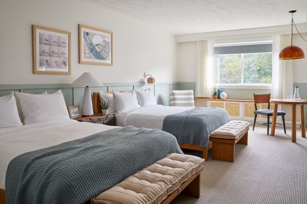 A cozy hotel room with two beds, a small table, chairs, decorative artworks, and a window with a view of trees outside, well-lit and inviting.