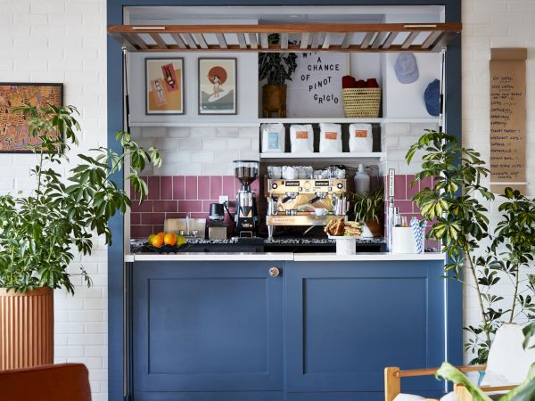 A small blue cafe counter with a pastry case, framed by plants, pink tile back wall, and cozy seating in a bright, airy nook.