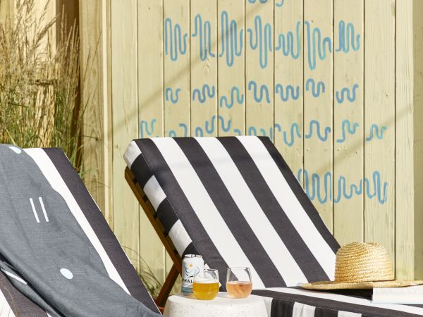 A striped outdoor lounge area with black-and-white chairs, a wooden privacy fence, a painted blue mural, and a small snack setup on a sunny day.