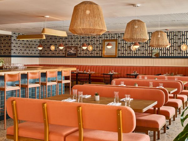 A retro diner interior with peach/orange booths, long wooden tables, tiled bar, wicker pendant lights, and a patterned wall backdrop.