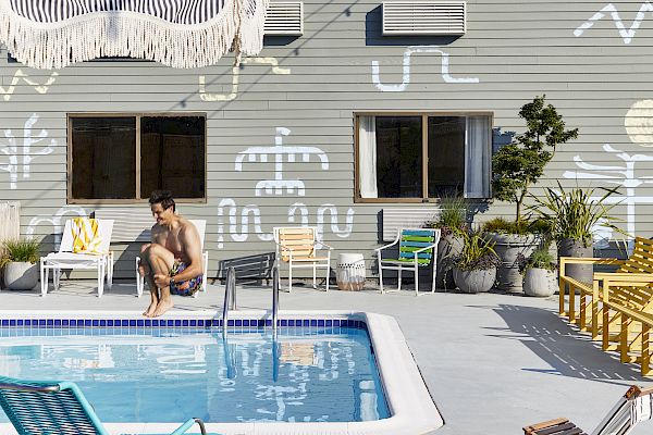 A poolside scene with people relaxing, colorful chairs, a wall with abstract art, and string lights against a clear sky.