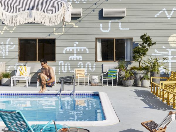 A poolside scene with people relaxing, colorful chairs, a wall with abstract art, and string lights against a clear sky.