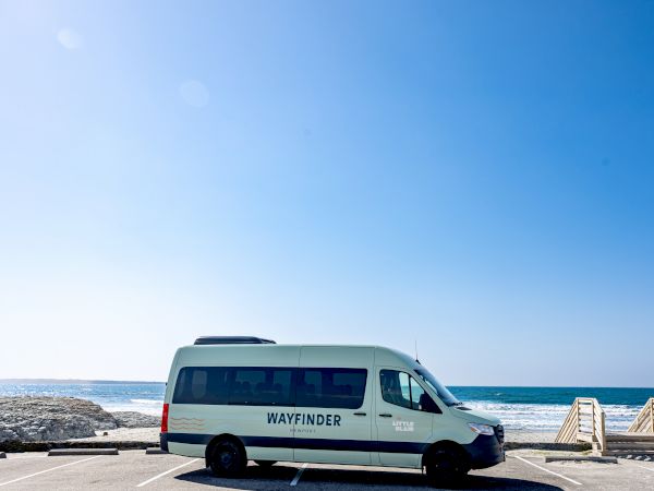 A white van marked "WAYFINDER" parked by a seaside promenade with a clear blue sky, calm ocean, and wooden steps to the beach.