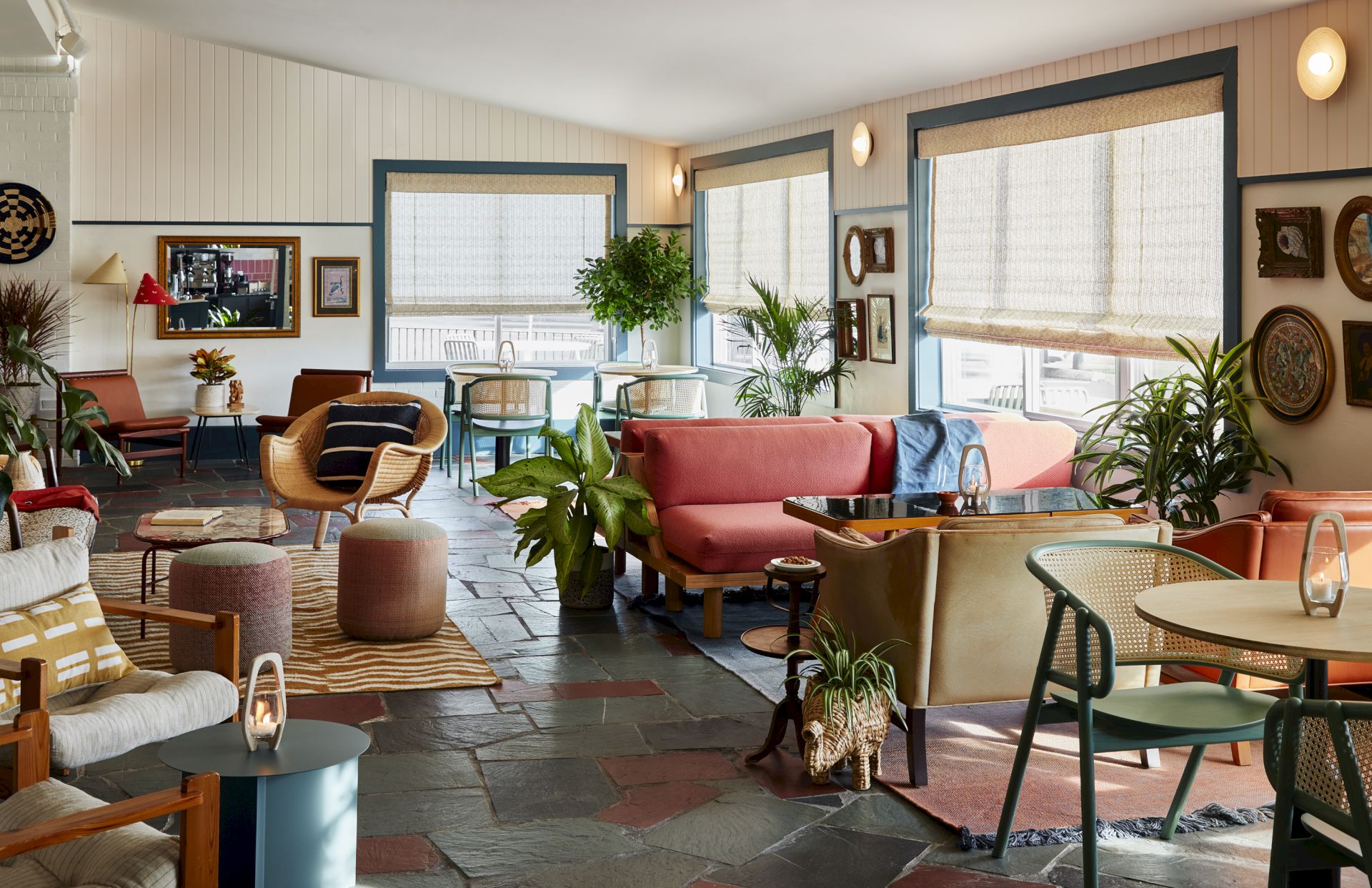 A cozy, stylish lounge with colorful sofas, chairs, plants, and framed art on the walls, filled with natural light from large windows.