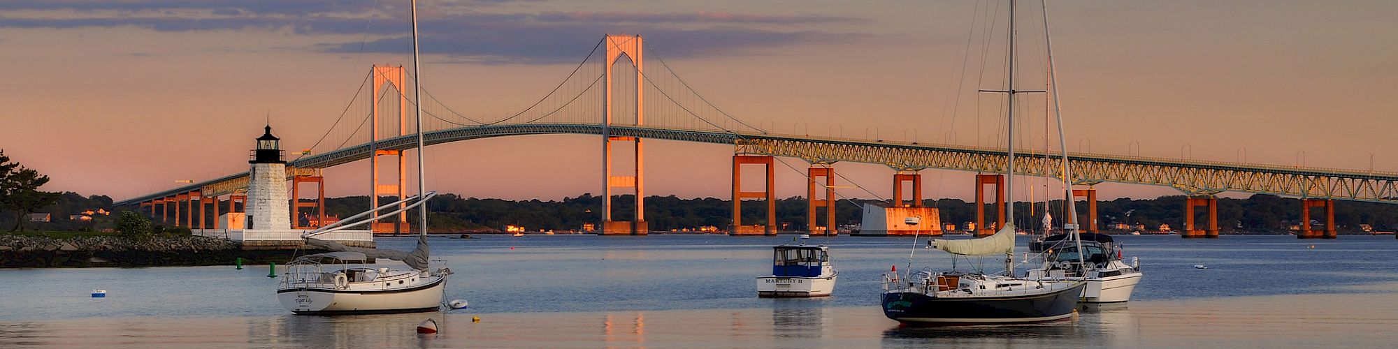 A scenic view of sailboats on calm water with a bridge and lighthouse in the background during a sunset.