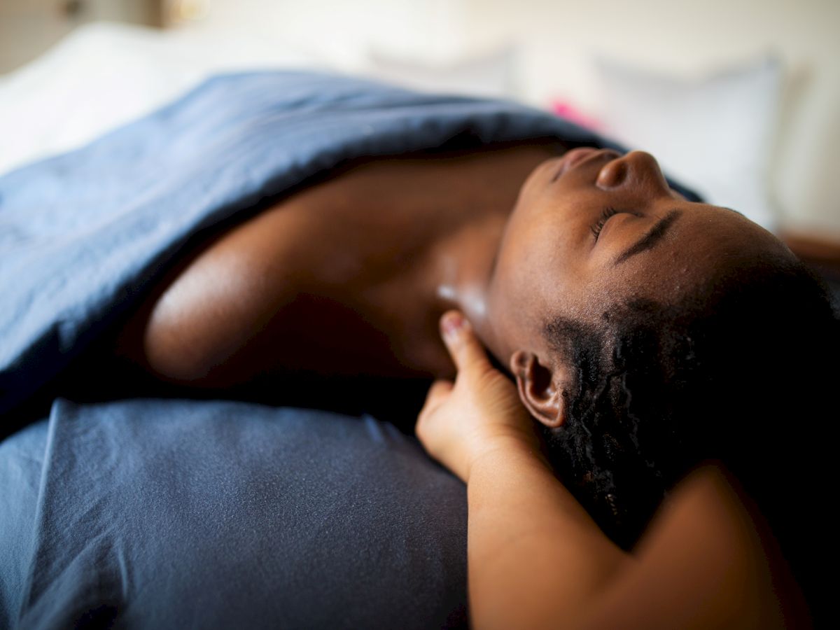 A person is lying under a dark blue blanket, receiving a shoulder massage in a relaxing setting.