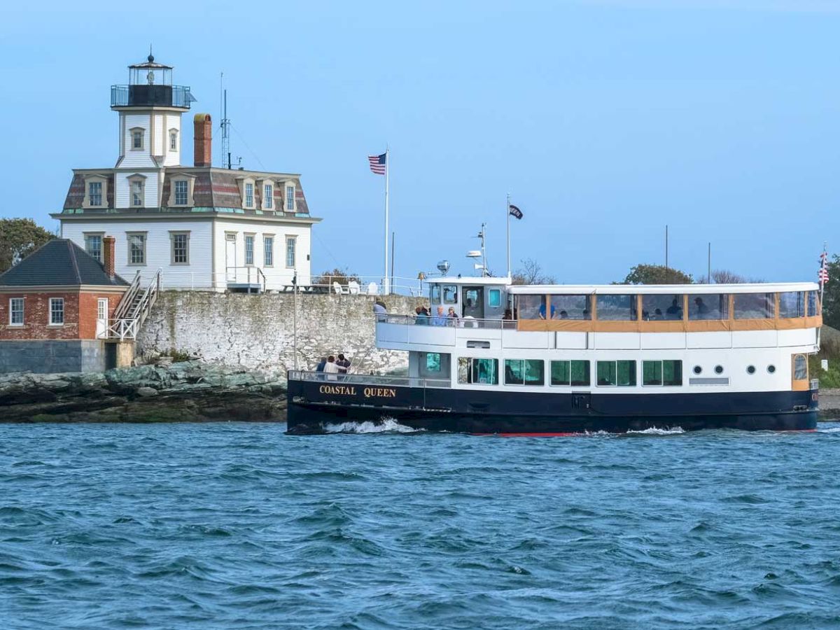 A lighthouse stands on the shore with a two-level ferry boat passing by on the water.