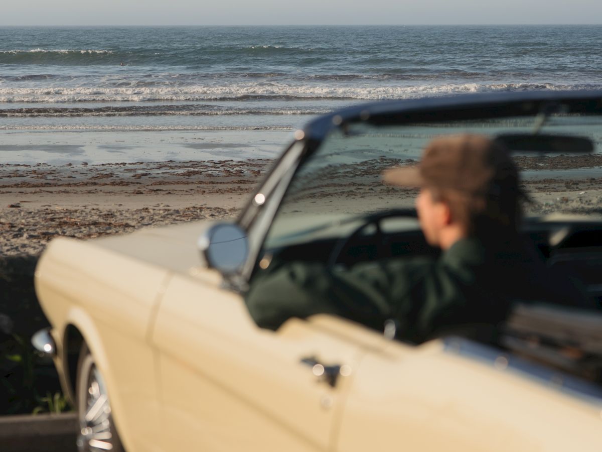 A white convertible is parked on a sandy beach as waves roll in under a cloudy sky, with a driver visible in the foreground.