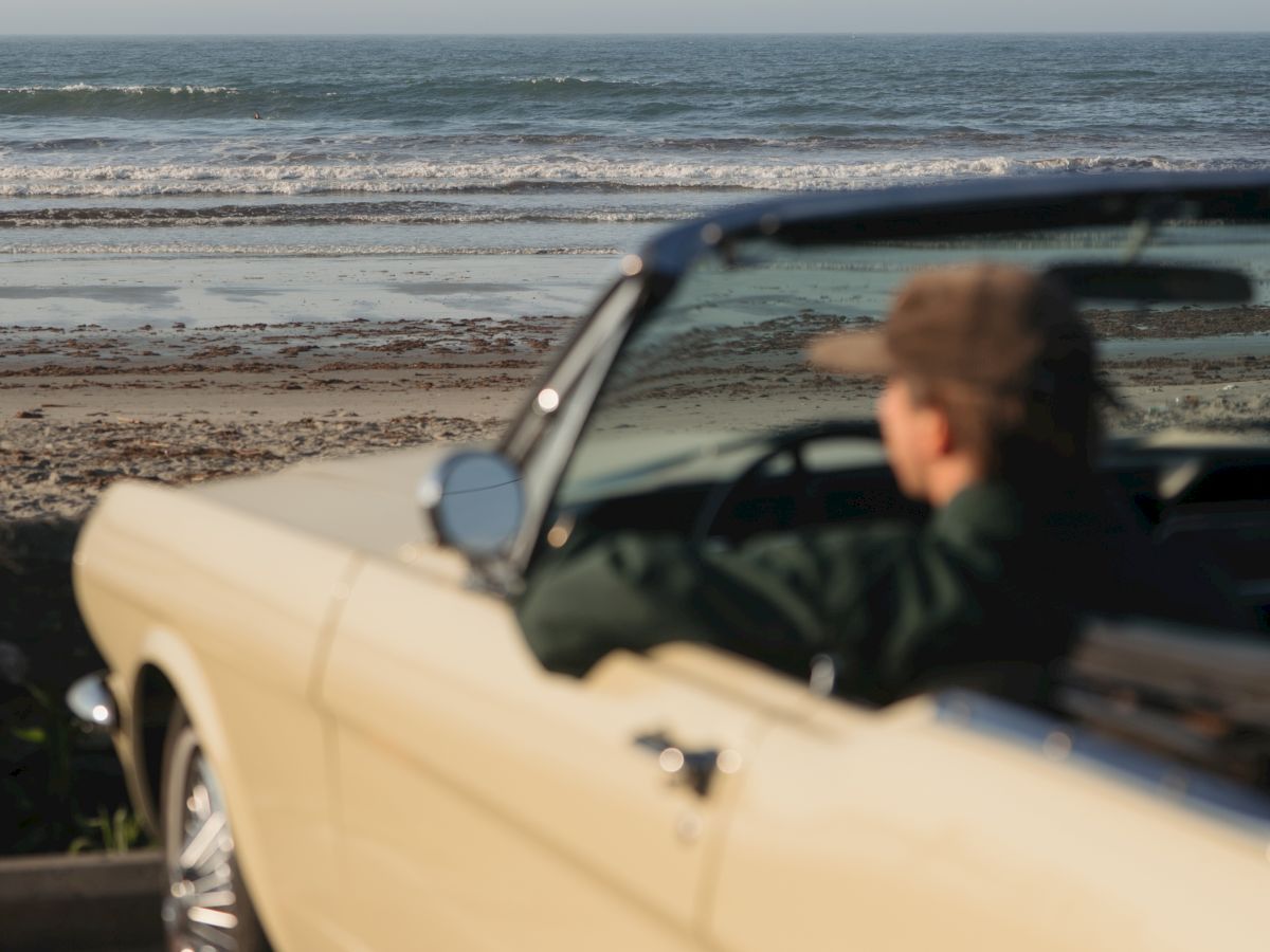 A white convertible parked on a sandy beach, with an ocean horizon and waves under a cloudy sky.