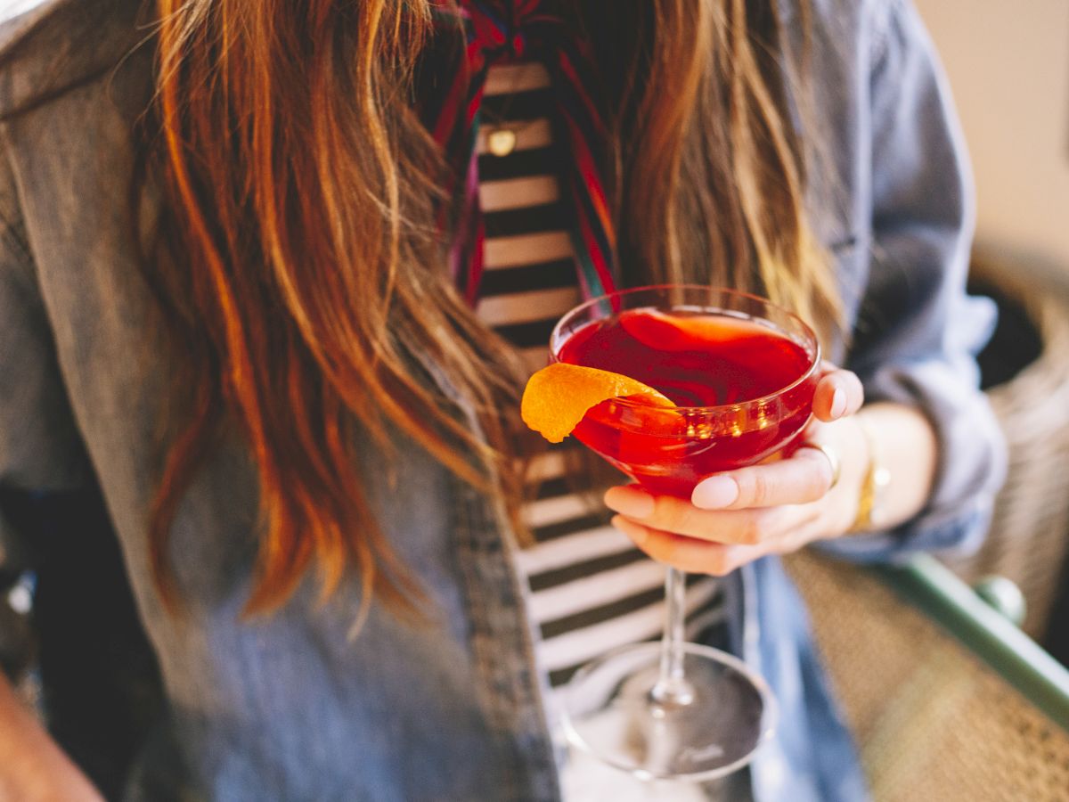 A person holding a red cocktail with an orange twist in a glass, wearing a striped shirt and denim jacket.