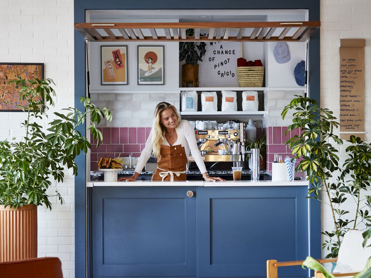 A small, cozy cafe with a blue counter, a woman behind it, plants, colorful tiles, and art on the walls.