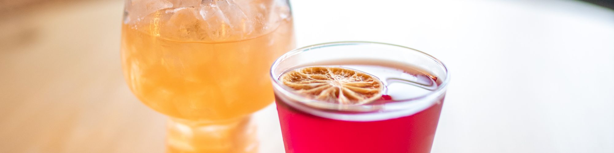 The image shows two colorful drinks on a table: one yellow-orange with a straw and garnish, and one pink with a citrus slice on top.