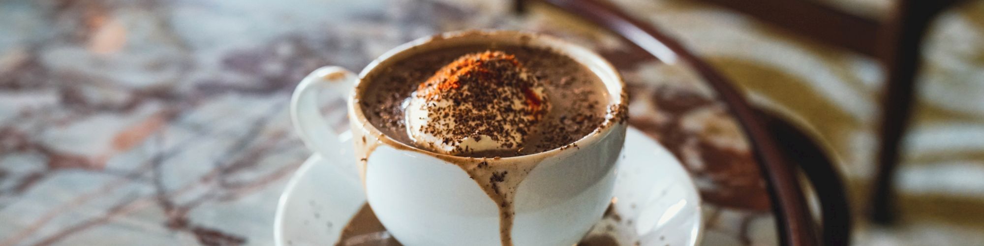 A cup of hot chocolate topped with whipped cream and chocolate shavings on a saucer, sitting on a marble table.