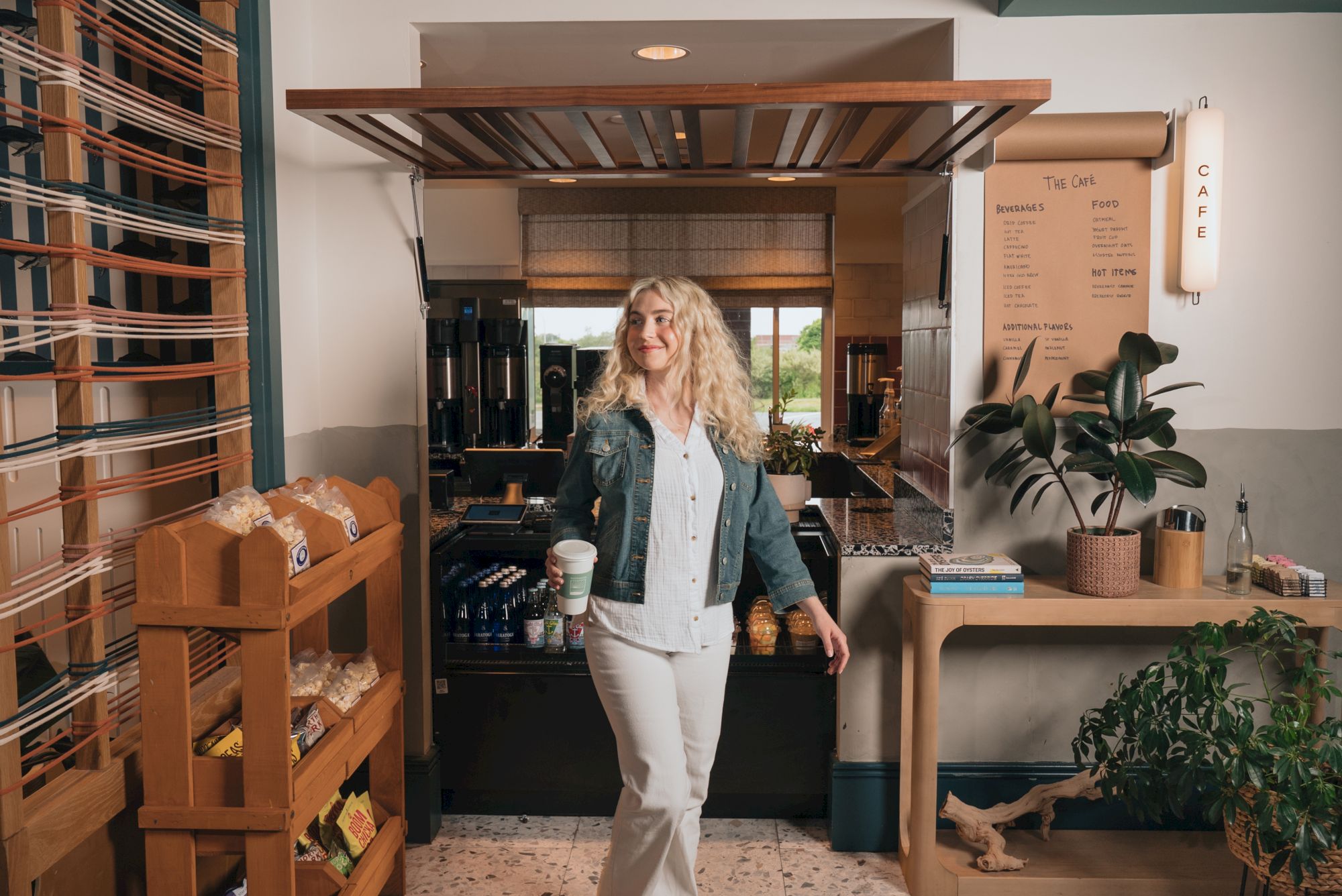 A blonde woman stands in a cozy caf&eacute; holding a coffee cup, with espresso machines, plants, and a &ldquo;The Cafe&rdquo; menu board behind her.
