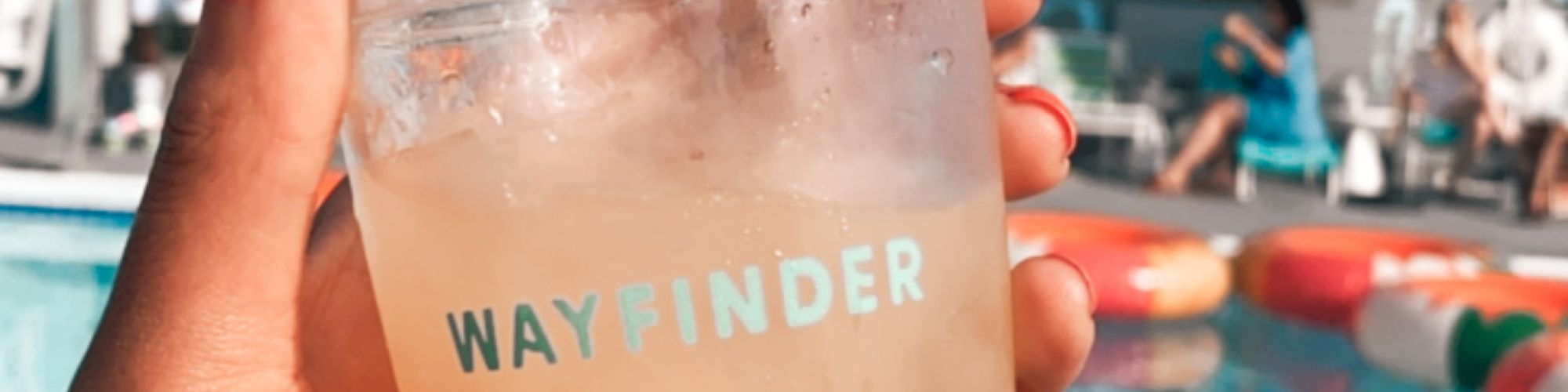 A hand holds a drink shaker labeled “WAYFINDER” in front of a pool and a building with abstract wall art.