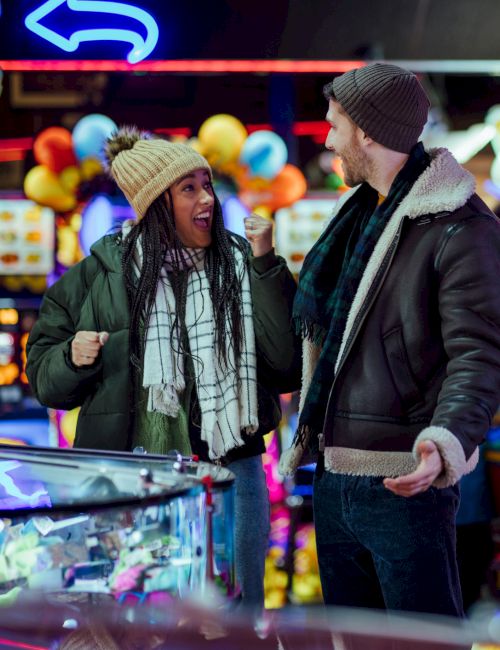 A man and woman, wearing winter clothes, are enjoying themselves in a colorful arcade filled with games and bright lights.