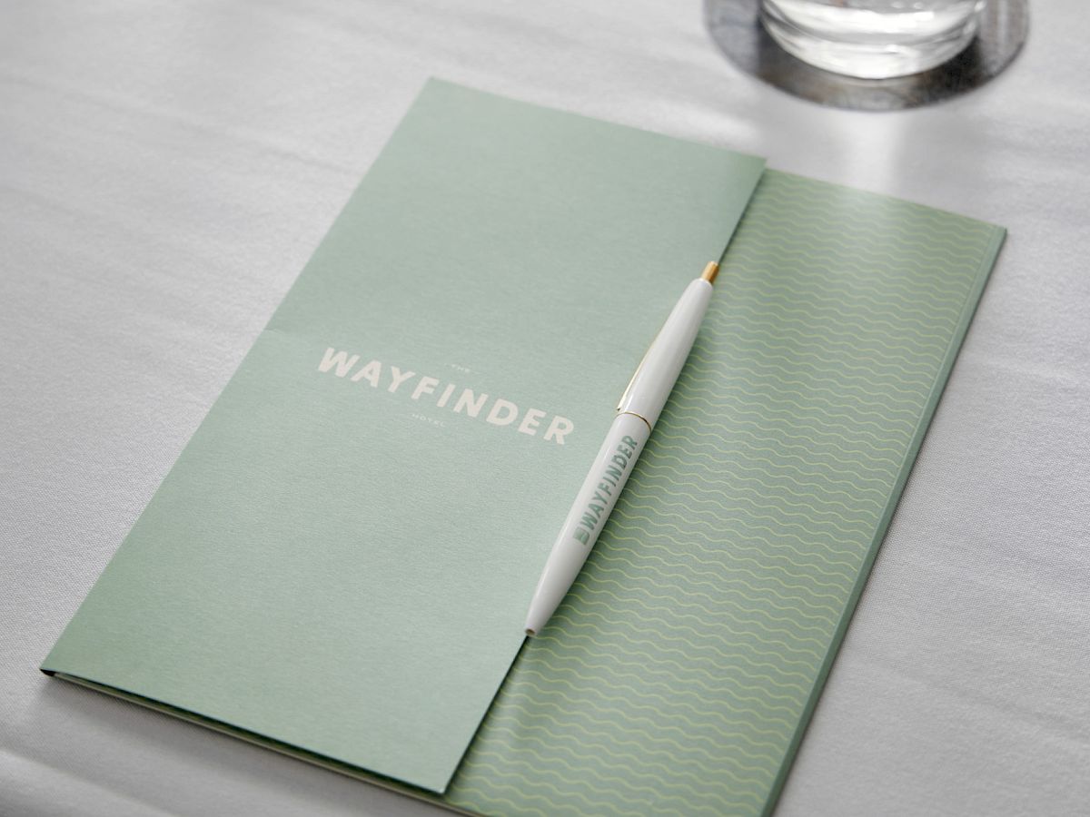 A notebook with the word "WAYFINDER," a pen, a glass of water on a coaster, and a bottle of water on a table.