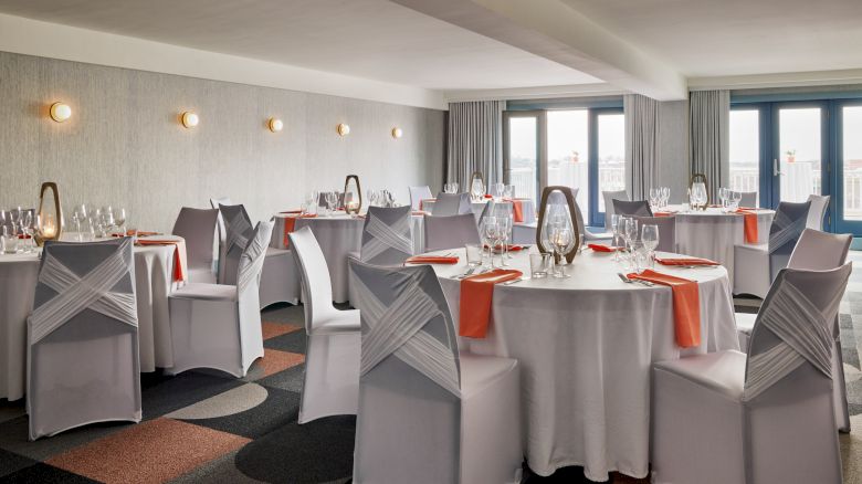 The image shows a banquet room with round tables set for an event, featuring chair covers and orange napkins on white tablecloths.