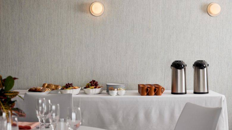 A table set for dining, with a buffet including pastries, fruits, and beverages, against a light-colored wall with two lights.