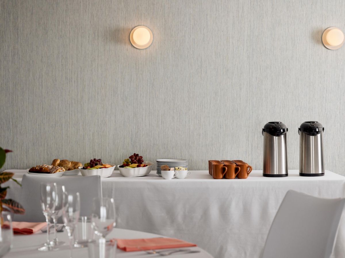 A table set with coffee urns, cups, and bowls of fruit and pastries; a dining table is in the foreground, set with glassware.