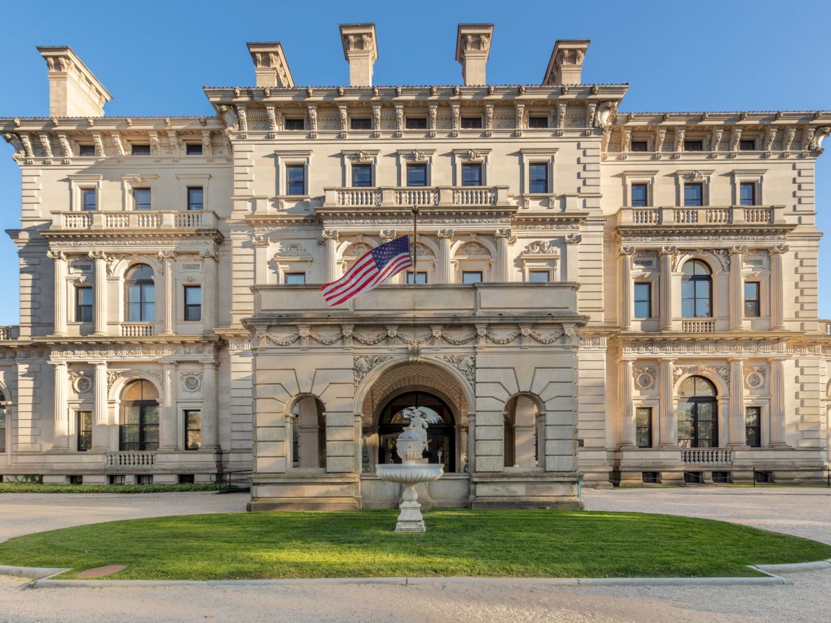 A grand, historic mansion with intricate architecture and a central archway, featuring an American flag at the front entrance.