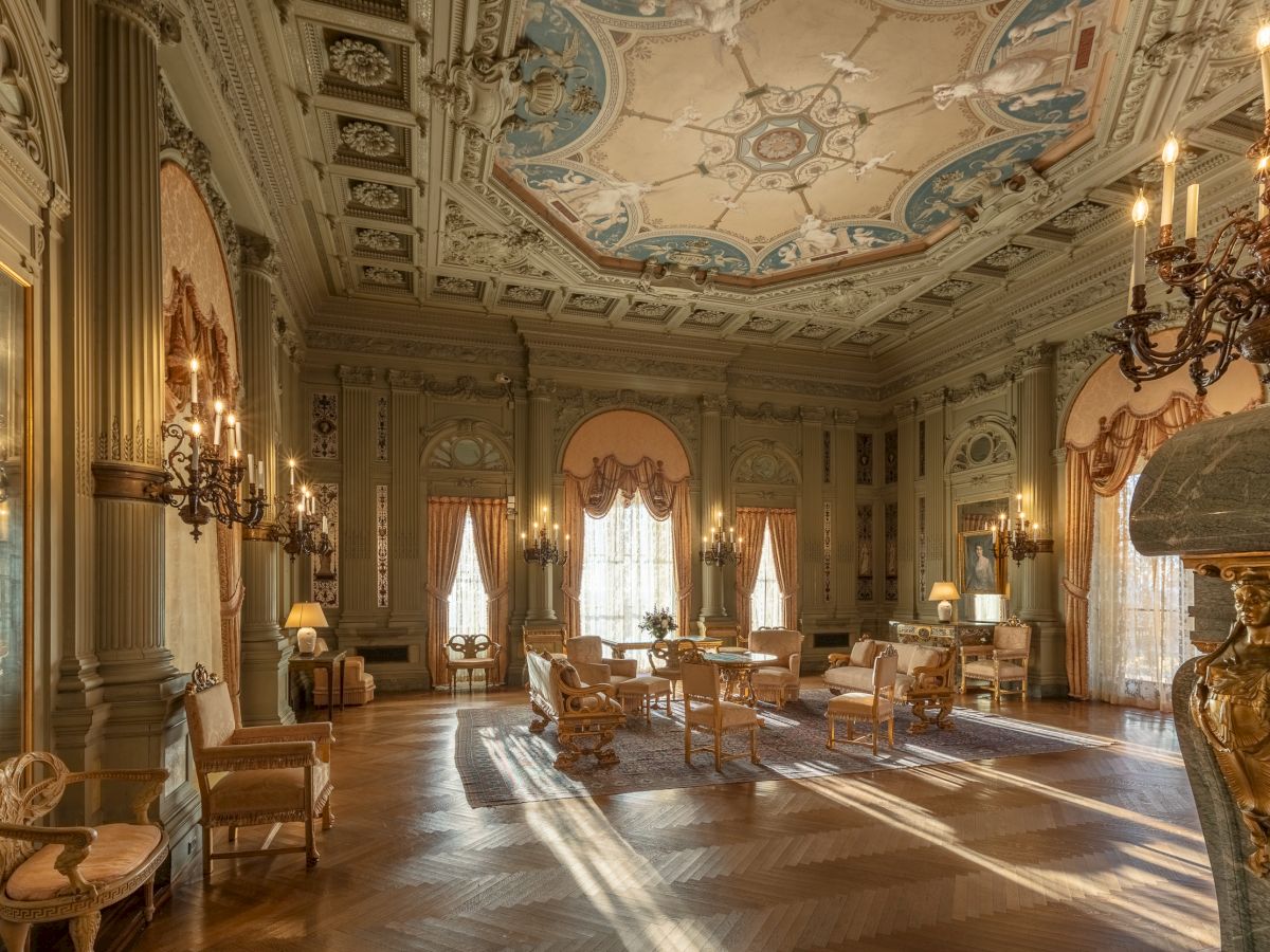 Luxurious antique lounge with ornate ceiling, chandeliers, gilded walls, and elegant seating bathed in sunlight streaming through tall windows.