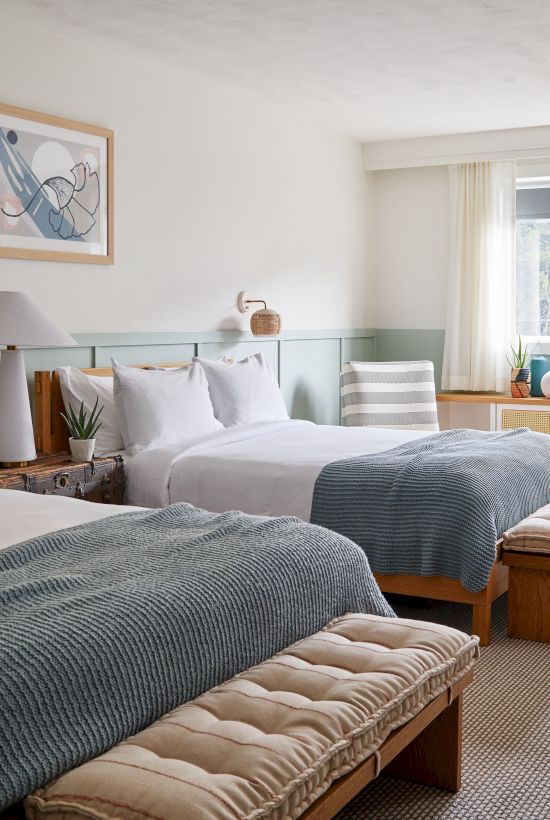 The image shows a cozy hotel room with two beds, art on the walls, a round table, and a window with natural light filtering in.