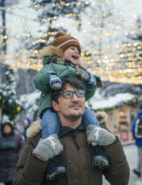 A man carries a child on his shoulders at a festive outdoor market with string lights.