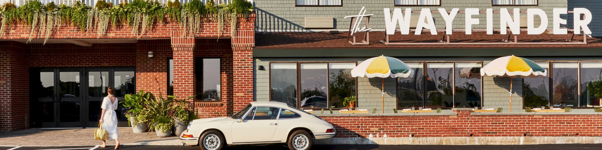 A vintage car is parked outside a building with "WAYFINDER" signage. A person walks nearby, and colorful umbrellas are visible.