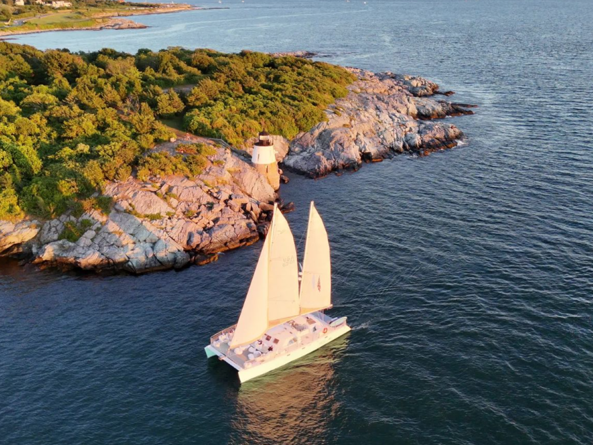 A white-sailed catamaran glides past rocky, tree-covered coastline as the sunlit water sparkles nearby.