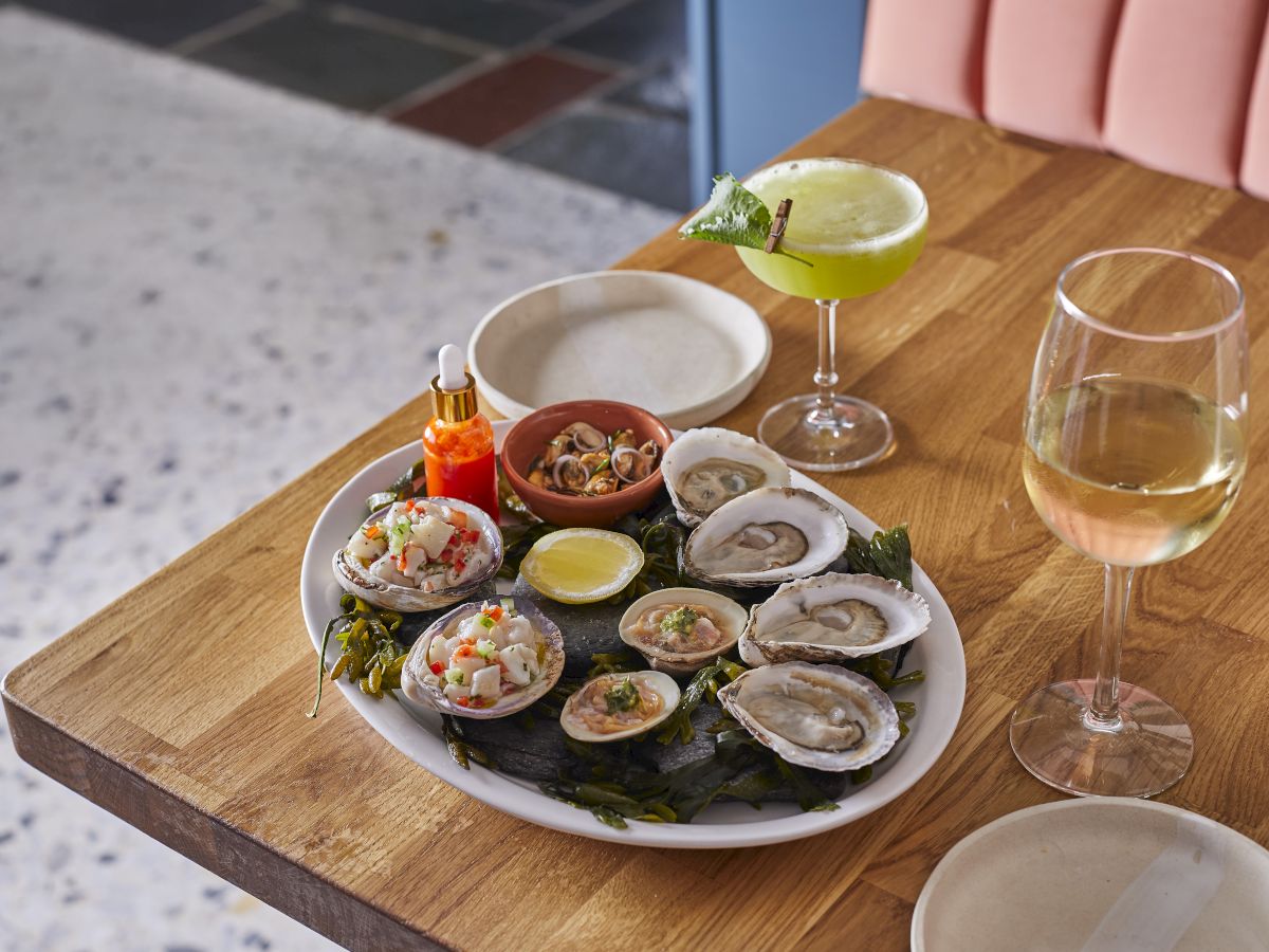 A seafood platter with oysters and clams, a green cocktail, and a glass of white wine are on a wooden table.