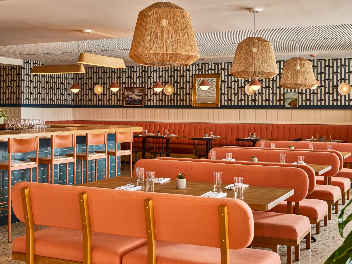 A retro-style diner with orange booths, wooden chairs, hanging wicker lights, a long bar, and a patterned wall behind the rows of tables.