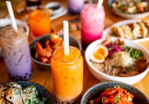 The image shows an assortment of colorful bubble teas and various dishes on a wooden table, featuring salads, bao buns, and fried chicken.