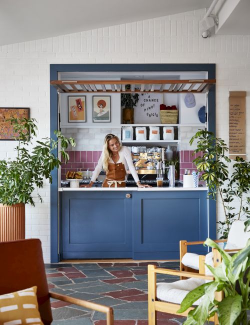 A person stands at a stylish blue coffee bar surrounded by plants and art, giving the space a cozy and inviting atmosphere.