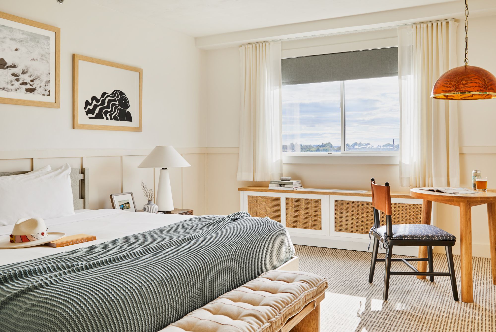 A cozy bedroom with a bed, wall art, a table with a chair, and a window with a scenic view, creating a relaxed atmosphere.