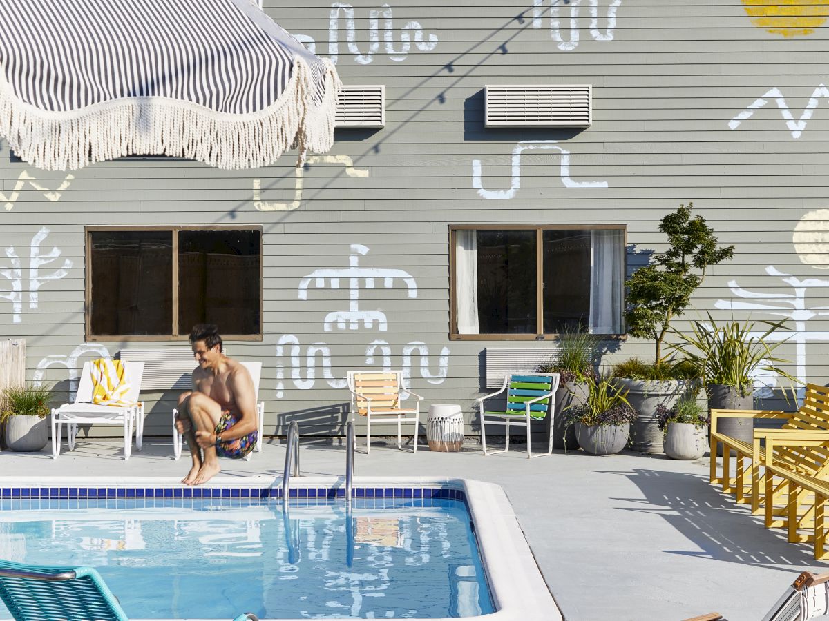A person by a pool, abstract wall art, string lights, chairs, umbrella, and plants create a summery outdoor scene.