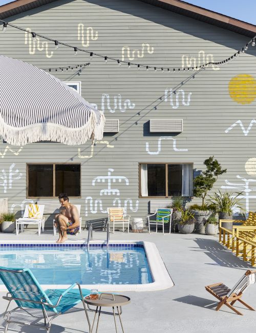 A person by a pool, abstract wall art, string lights, chairs, umbrella, and plants create a summery outdoor scene.
