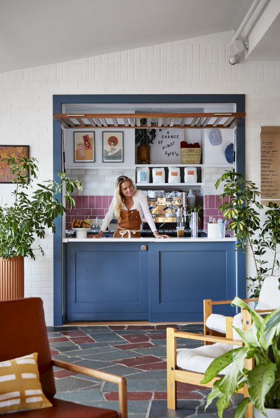 A person stands at a stylish blue coffee bar surrounded by plants and art, giving the space a cozy and inviting atmosphere.