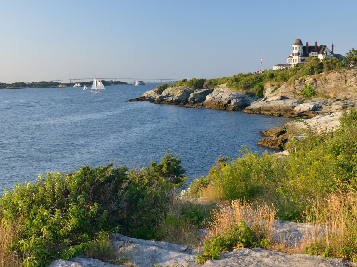 A coastal landscape with a house on a cliff, sailboats in the water, and greenery in the foreground.