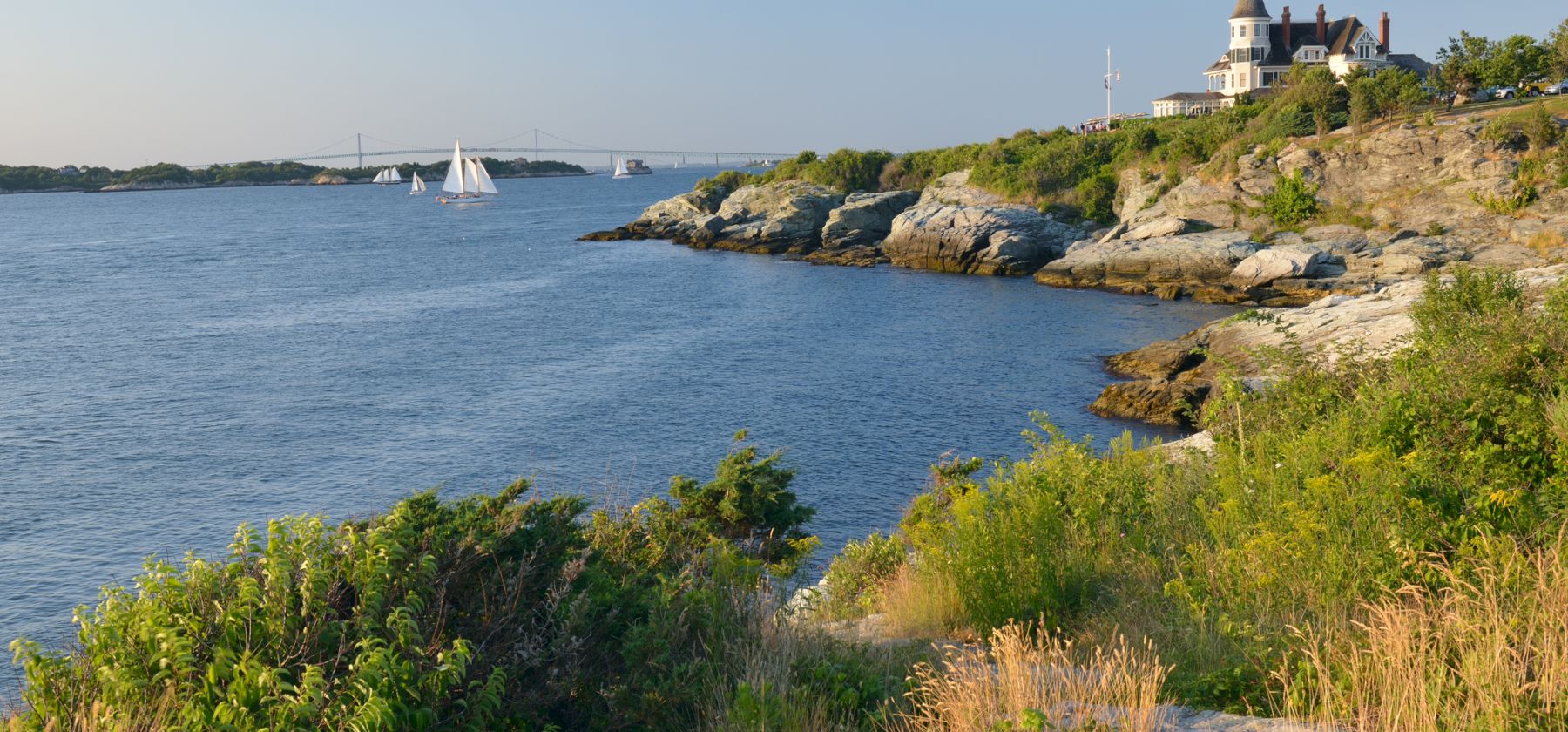 A coastal landscape with a house on a cliff, sailboats in the water, and greenery in the foreground.