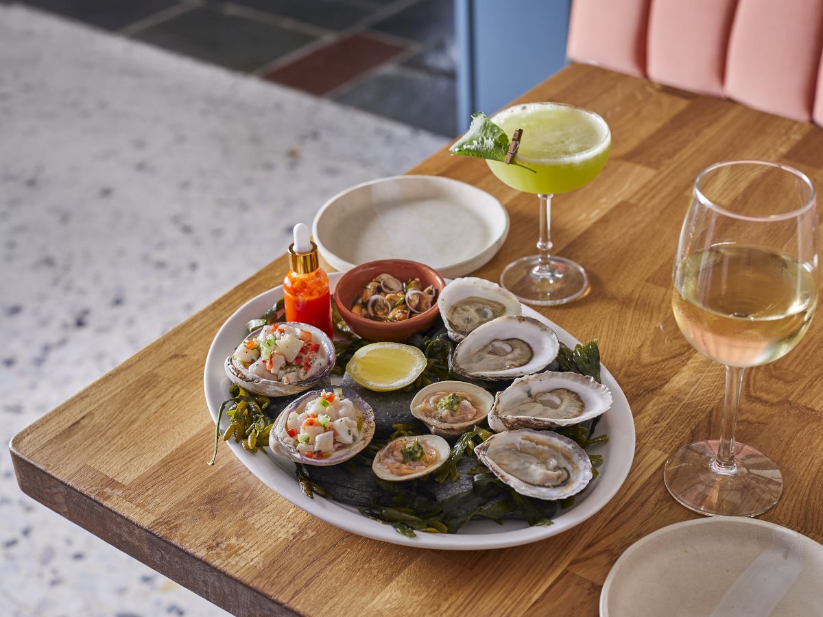 A plate of oysters and shellfish with sauce and lemon wedges, accompanied by a green cocktail and a glass of white wine.