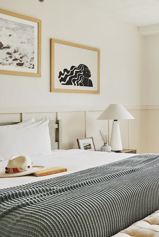 This image shows a modern, well-lit bedroom with a neatly made bed, artwork on the walls, a window with a cityscape view, and a small study table.