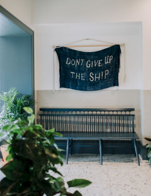 A room with a banner saying "DON'T GIVE UP THE SHIP," a bench below, and plants nearby.