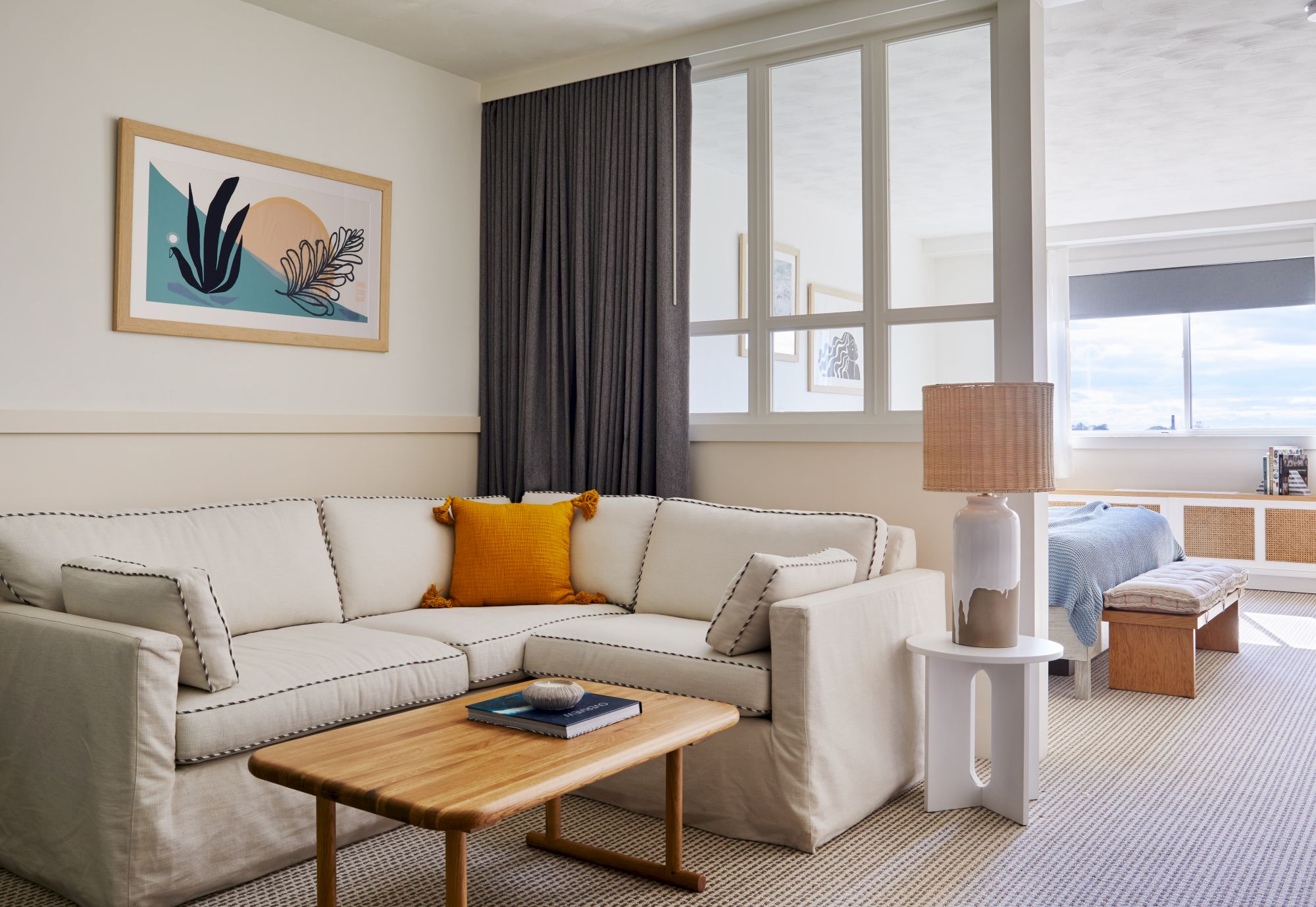 A cozy living room with a beige sectional sofa, yellow cushion, wooden coffee table, framed art, lamp, and a glimpse into a sunlit bedroom.
