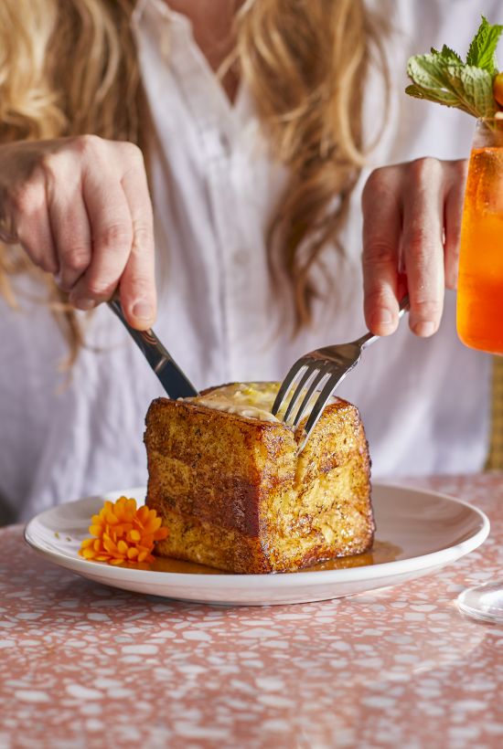 A person cuts into a thick slice of French toast on a plate with a flower garnish, next to a vibrant orange drink in a stemmed glass.