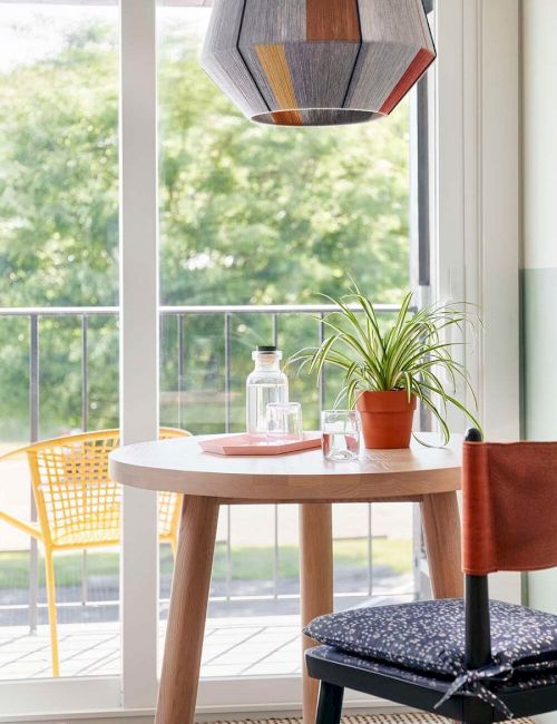 A cozy dining area with a wooden table, plant, glassware, colorful lamp, chair, and balcony view with wicker chair.