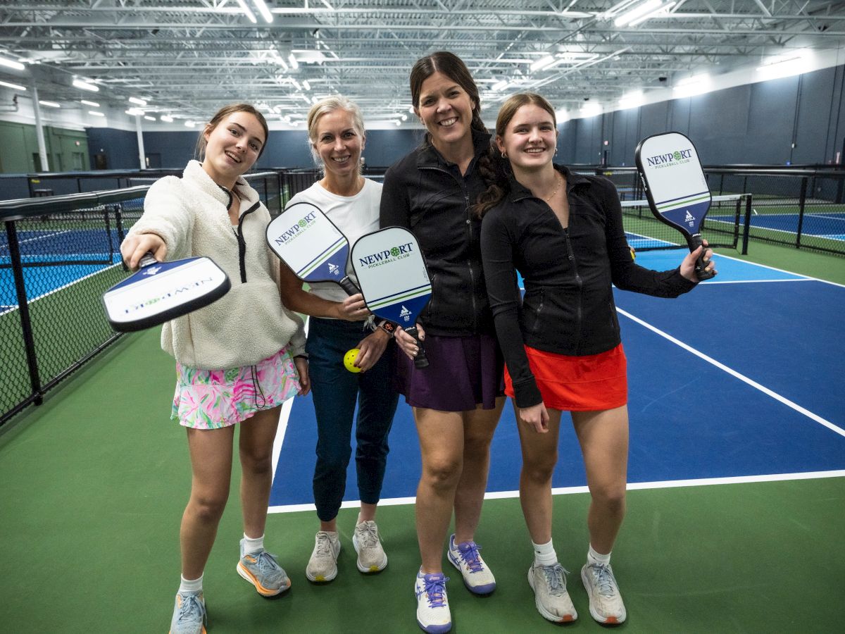 Four people holding pickleball paddles stand on an indoor court, smiling at the camera.