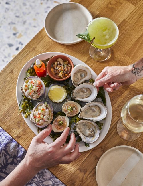 A seafood platter with oysters, ceviche, lemon wedges, a small bottle of hot sauce, and drinks on a wooden table.